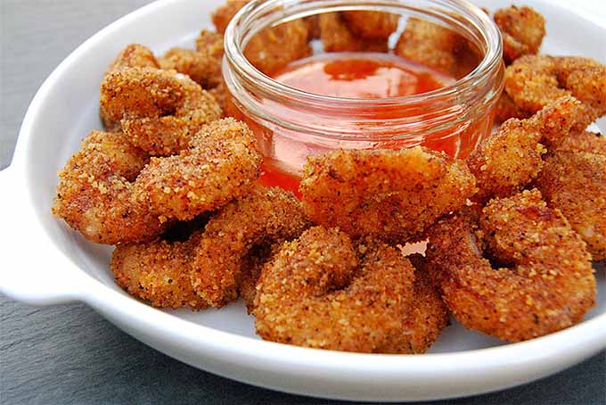Breaded prawns on a platter surrounding a clear glass jar of red dipping sauce.