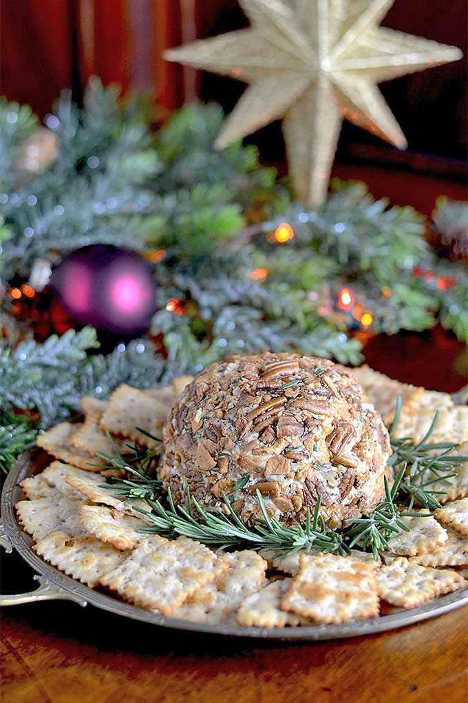 A cheese ball encrusted with nuts and herbs in the foreground on a plate surrounded by crackers and rosemary, with evergreen boughs, ornaments, and decorative lights in the background.