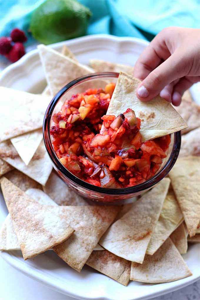 A hand dips a chip into a glass bowl of fruit salsa surrounded by more triangular chips, with a lime and raspberries in the background on a blue cloth napkin.
