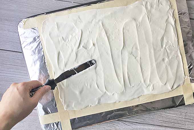 A human hand spreads a layer of white chocolate on a layer of aluminum foil attached to a baking sheet.