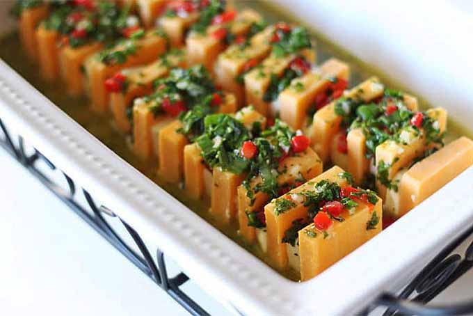 Sliced marinated cheese arranged in rows in a ceramic serving dish and topped with finely chopped herbs.