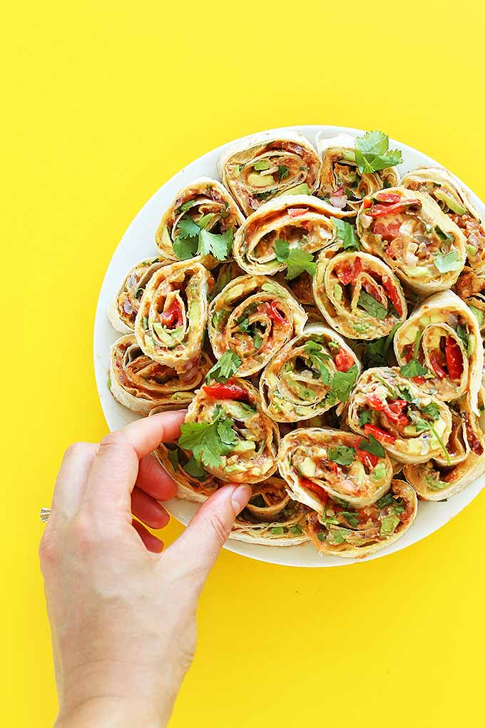 Pinwheel wraps stuffed with vegetables and vegan cheese arranged on a white plate with a hand reaching for one, on a bright yellow background.
