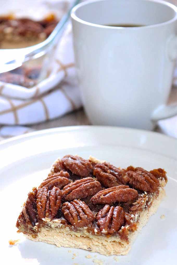 A pecan pie shortbread bar on a plate, with more in the background in a clear glass baking dish, and a white ceramic mug of coffee.