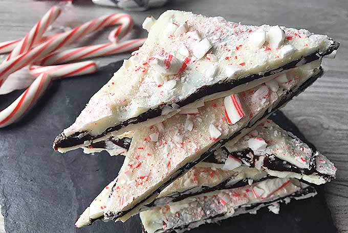Close up of a small pile of homemade Christmas white and dark chocolate peppermint bark candy sitting on a black slate surface. The bark is positioned so the viewer can see the layers.