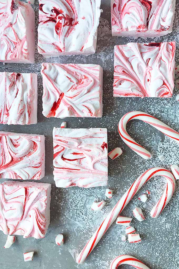 Top down view of squares of homemade peppermint marshmallows lined up on a gray slate surface. Two candy canes are to the lower right.