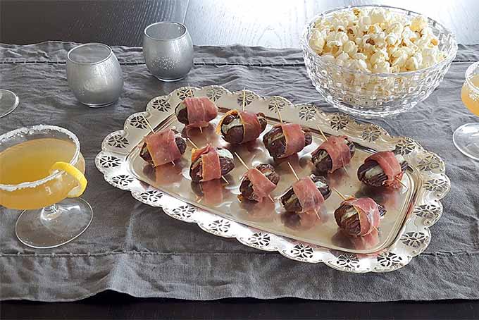 Dates stuffed with blue cheese and wrapped in thinly sliced deli meat, arranged in two rows on a sterling silver tray with a lemon cocktail to the left and two silver votive candle holders, and a glass bowl of popcorn to the right, on a gray wrinkled tablecloth.