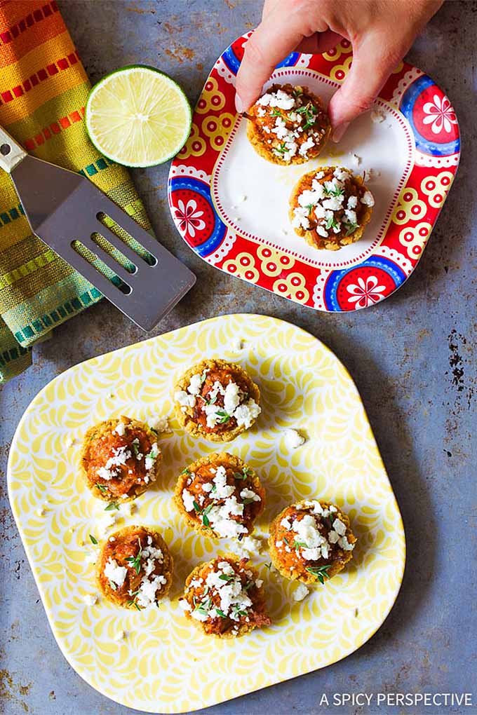 Overhead closely cropped image of tamale bites on two patterned plates, topped with cotija cheese and herbs, with a metal spatula, half of a lime, and a rainbow-colored striped cloth napkin.