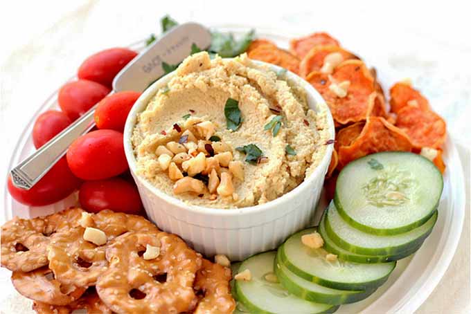 A large ceramic remekin of vegan cheese topped with nuts and herbs, on a platter surrounded by pretzels, chips, sliced cucumber, and grape tomatoes, with a silver serving spreader.
