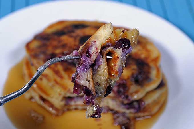 A close up of a fork with a bite of blueberry butter milk pancakes with the remainder of the stack in a diffused background | Foodal