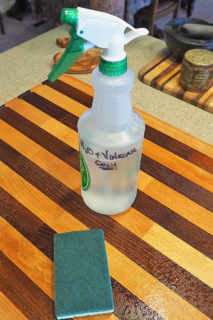 A bottle of 50% vinegar and 50% water is sitting on a wooden cutting board along with a a green scratch pad/sponge combo | Foodal