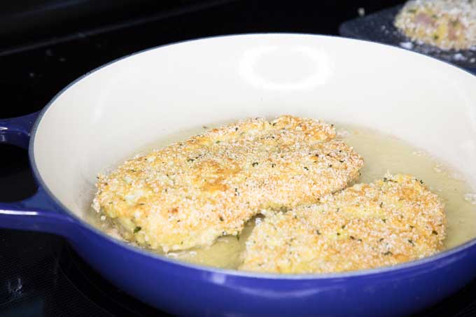 Oblique view of breaded chicken breasts inside of a porcelain coated cast iron pan full of heated cooking oil | Foodal