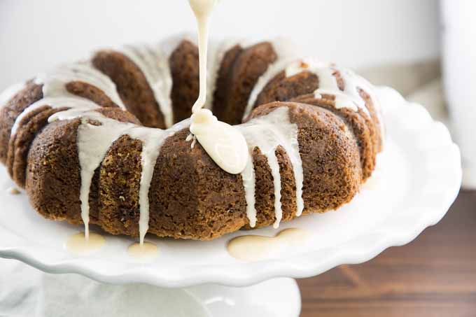 Square image of a brown banana bundt cake with white maple glaze being drizzled onto the top of the cake from above, on a white cake stand atop a brown wooden table.