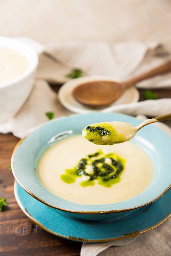 Vertical image of a golden spoon full of celery and apple soup topped with chive oil being held up to the camera, with a blue bowl with saucer of the soup in the background, next to a wooden serving spoon on a white spoon rest, a larger white bowl of soup, a white cloth, and scattered fresh herbs on a brown wooden tabletop.