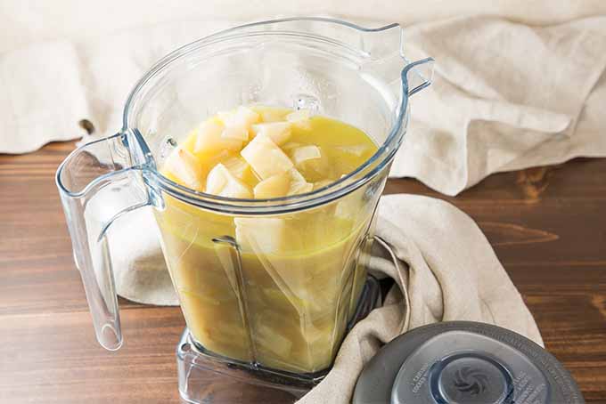 A plastic blender canister filled with a yellow broth, and chopped apples and celery root, on a brown wooden table wrapped in a white cloth, beside the blender lid.