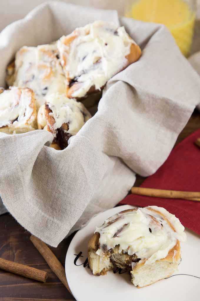 Vertical image of a cloth napkin-lined basket of homemade cinnamon buns topped with cream cheese glaze, with one bun on a small white plate to the right, on a brown wooden table topped with a red cloth, with scattered cinnamon sticks.
