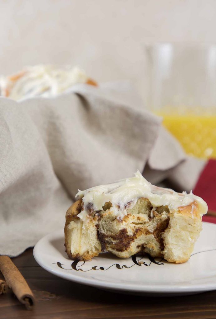 Vertical image of a partially eaten cinnamon roll on a small white plate, with a gray cloth-lined basket of more cinnamon rolls and a glass of orange juice in the background.