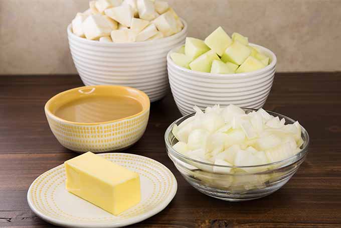 Ingredients for making a soup- a white plate of butter, a bowl of broth, and bowls of chopped apples, celery root, and onions, on a brown wooden surface with a beige background.