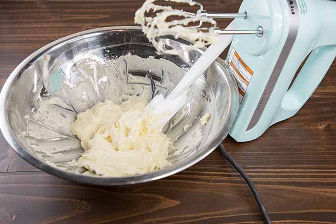 A stainless steel bowlful of cream cheese glaze for topping cinnamon rolls, with a white rubber spatula stuck into the icing, an a blue hand mixer with beaters coated in the mixture propped up to the right of the bowl on a brown wooden table.