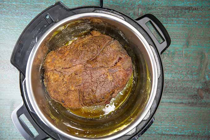 A seared whole chuck roast topped with some liquid in a slow cooker on a blue stained wooden surface.