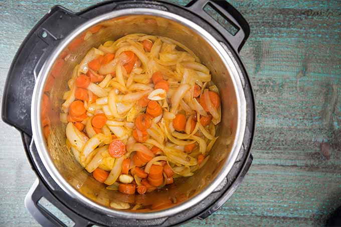 Cooked sliced onions and carrots in the bottom of a slow cooker, on a stained blue wooden surface.
