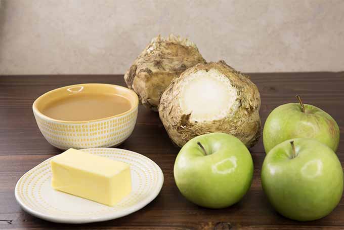 A small white plate of butter, a dish of broth, two whole celery roots and three green apples, on a brown wooden table with a beige wall in the background.