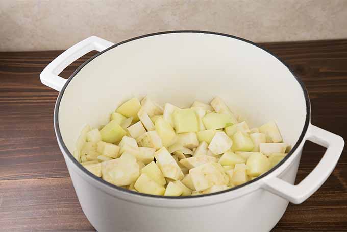 A white enamel pot filled with chopped celery root, apples, and onion, on a brown wooden surface with a beige background.