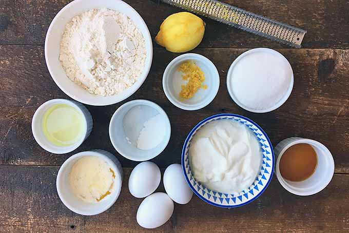 Horizontal image of ingredients in bowls, eggs, a lemon, and a zester on a wooden surface.