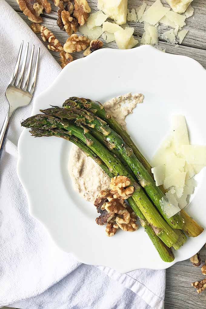 Vertical top-down image of a plate of asparagus surrounded by nuts and cheese on a white towel with a fork on a gray wooden surface.