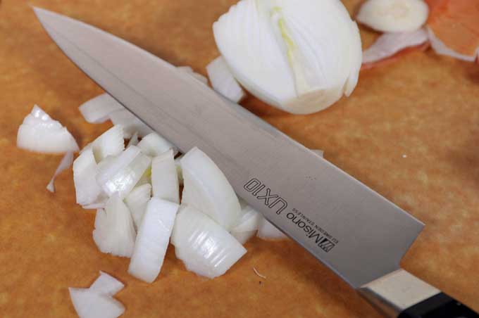 Onions being chopped with a Japanese chef's knife on a brown cutting board.