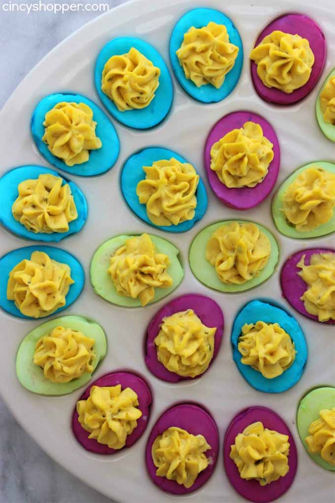 At least twenty deviled eggs with whites that have been dyed blue, green, and purple, arranged in concentric circles in a large egg serving dish with wells to hold the eggs, on a gray background.