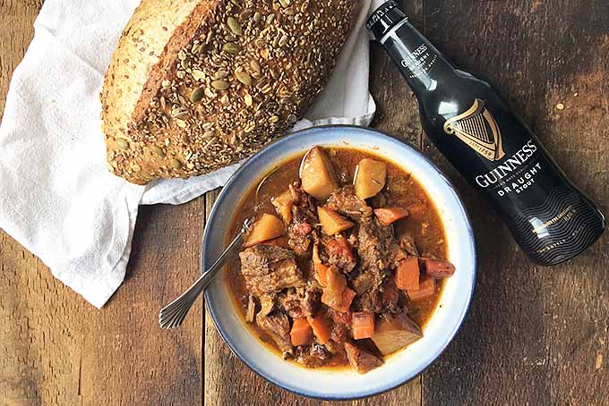 Horizontal image of a bowl of beef stew on a white towel with beer and bread on a wooden surface. 