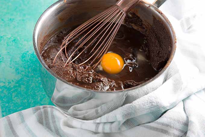 A stainless steel mixer bowl filled with chocolate brownie batter with an egg cracked on top, with a copper-colored whisk and a white and gray striped kitchen towel wrapped around the base of the bowl, on a robin's egg blue countertop.