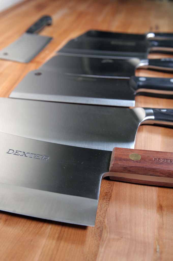 Oblique view of various meat cleavers on a wooden prep table; the foreground is in focus and the background is starting to become diffused.