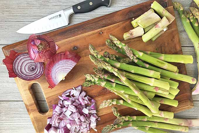 Horizontal image of a cutting board with chopped asparagus and red onions with a knife on a gray wooden surface.
