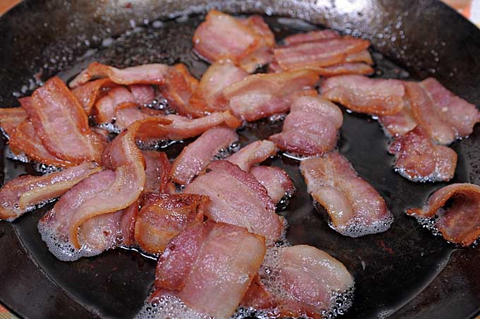 Close up of bacon pieces being fried in a carbon steel skillet.