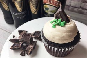 Horizontal image of a brown cupcake topped with white buttercream and chocolate/green sprinkle garnishes on a white plate with more garnishes, and bottles in the background.