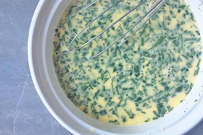 Horizontal image of a whisk in a bowl with a parsley and egg mixture on a gray surface.