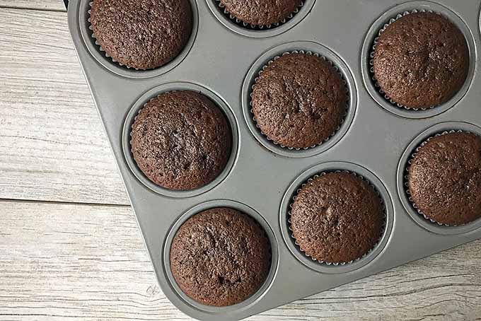Horizontal top-down image of baked brown cupcakes in muffin trays on a gray wooden surface. 
