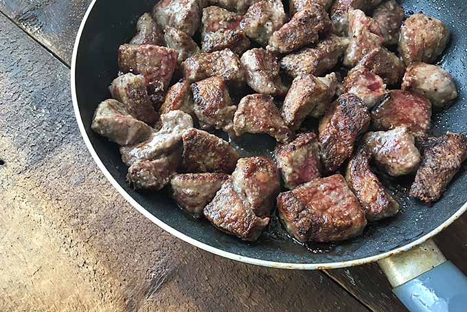 Horizontal image of browned steak pieces in a skillet on a wooden surface. 