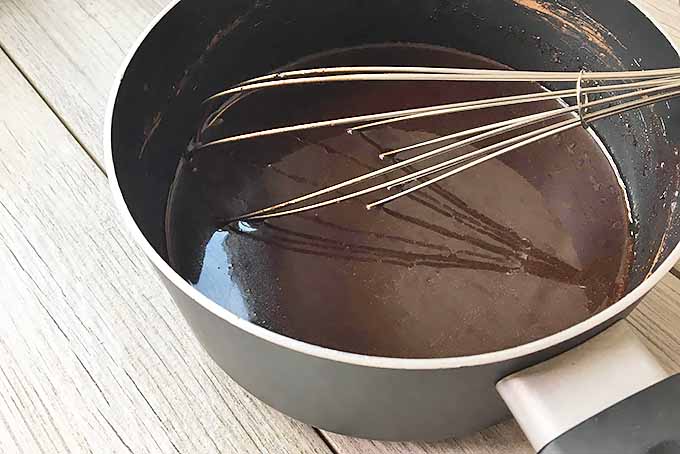 Horizontal image of a metal whisk dipped in a dark brown liquid mixture in a saucepan on a gray wooden surface.