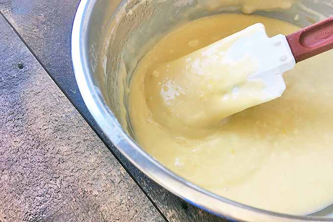 Horizontal image of a rubber spatula mixing a yellow batter in a metal bowl on a wooden surface.