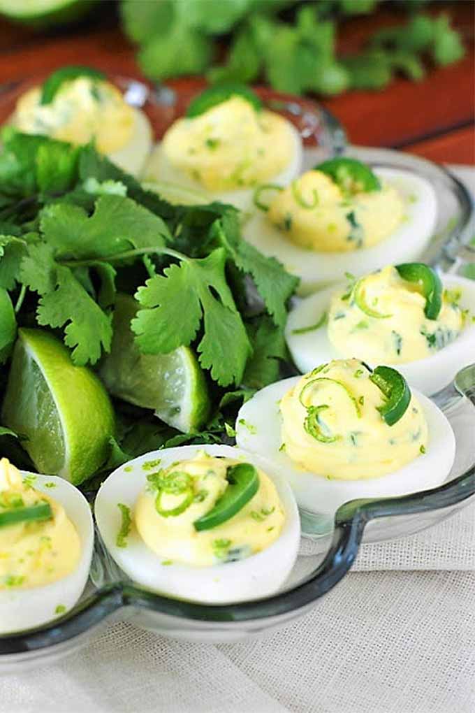 Vertical closeup of seven deviled eggs arranged on a glass scalloped egg serving dish, topped with chopped jalapeno and cilantro, with larger cilantro leaves and limewedges at the center of the bowl, and more cilantro in shallow focus in the background.