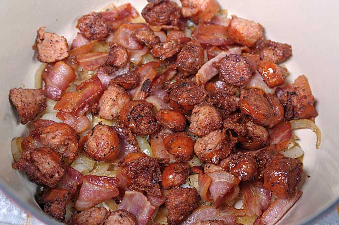 Close up of sausage pieces, bacon, and fried onion being layered into the bottom of red porcelain coated Dutch oven.