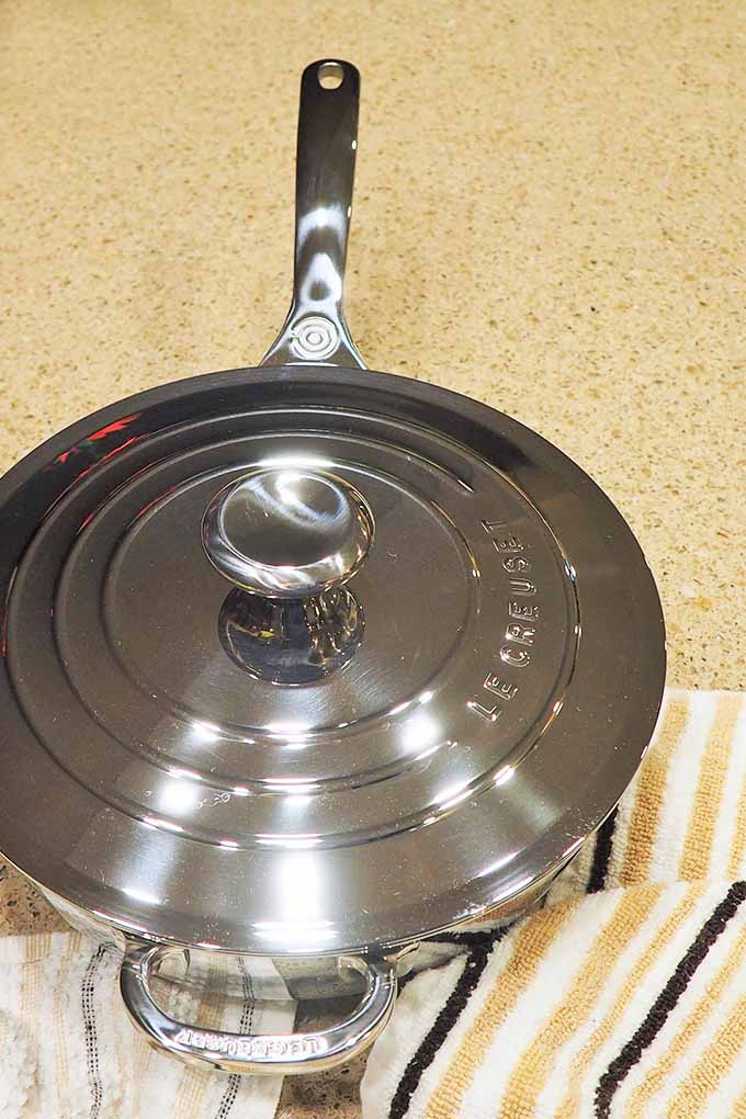 Top view of a stainless steel Le Creuset saucepan with lid, on a beige countertop.