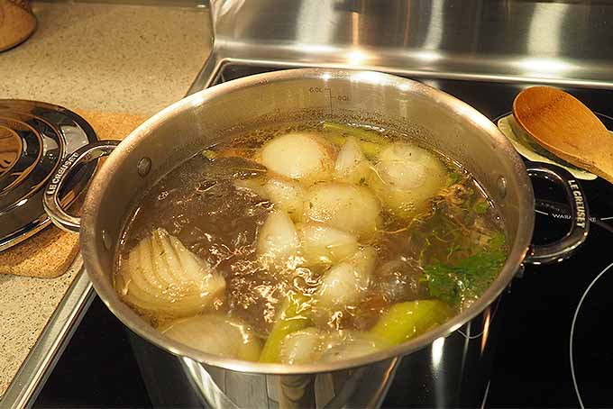 Top view of a large stainless steel stockpot filled with broth, vegetables, and herbs on a stove.