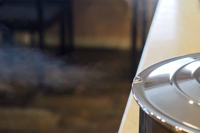Closeup of a stainless steel pan with a hole for steam venting in the top, with steam coming out of the vent, with a brown and beige kitchen in shallow focus in the background.