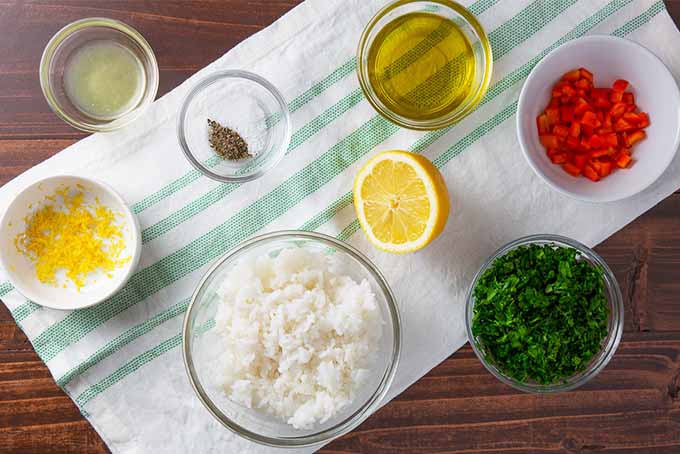 Overhead view of all of the ingredients needed to make a simple rice salad- small bowls of lemon zest, lemon juice, salt and pepper, olive oil, cooked rice, diced red bell pepper, minced parsley, and half of a lemon, all on a white and pale green striped kitchen towel on a brown wooden surface.