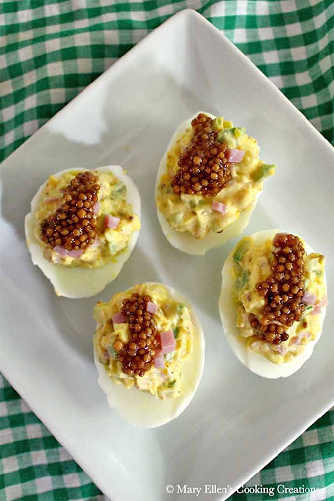 Top-down vertical image of four hard-boiled eggs on a plate, cut in half and filled with a yolk mixture, topped with pickled mustard seeds.