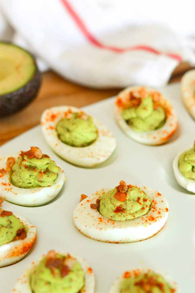 Hard boiled egg white halves filled with a green egg mixture topped with paprika and bacon, and arranged in the wells of a round pastel blue egg serving plate, on a wooden countertop with half of an avocado and a white dishcloth with a red stripe in the background.