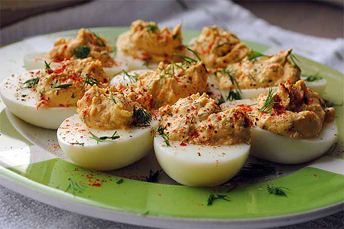 Nine halved hard-boiled egg whites filled with a creamed yolk mixture, topped with paprika and chopped herbs, on a green patterned plate with a cloth under it.